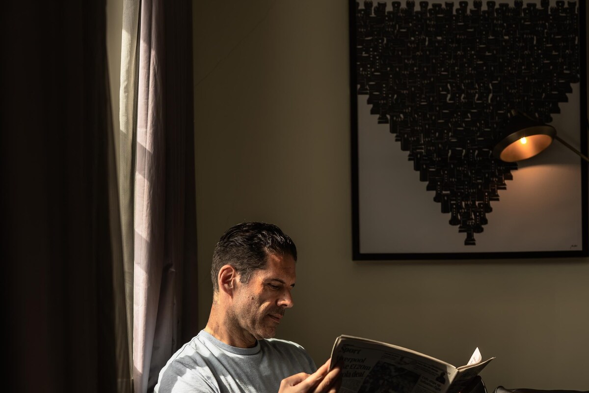 A person sitting on a chair reading a newspaper in a room with a lamp and a framed picture on the wall.