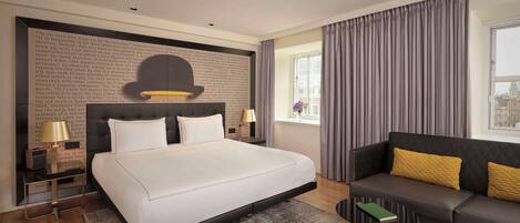 A modern hotel room with a large bed, a sofa, a small table, and a window with curtains.