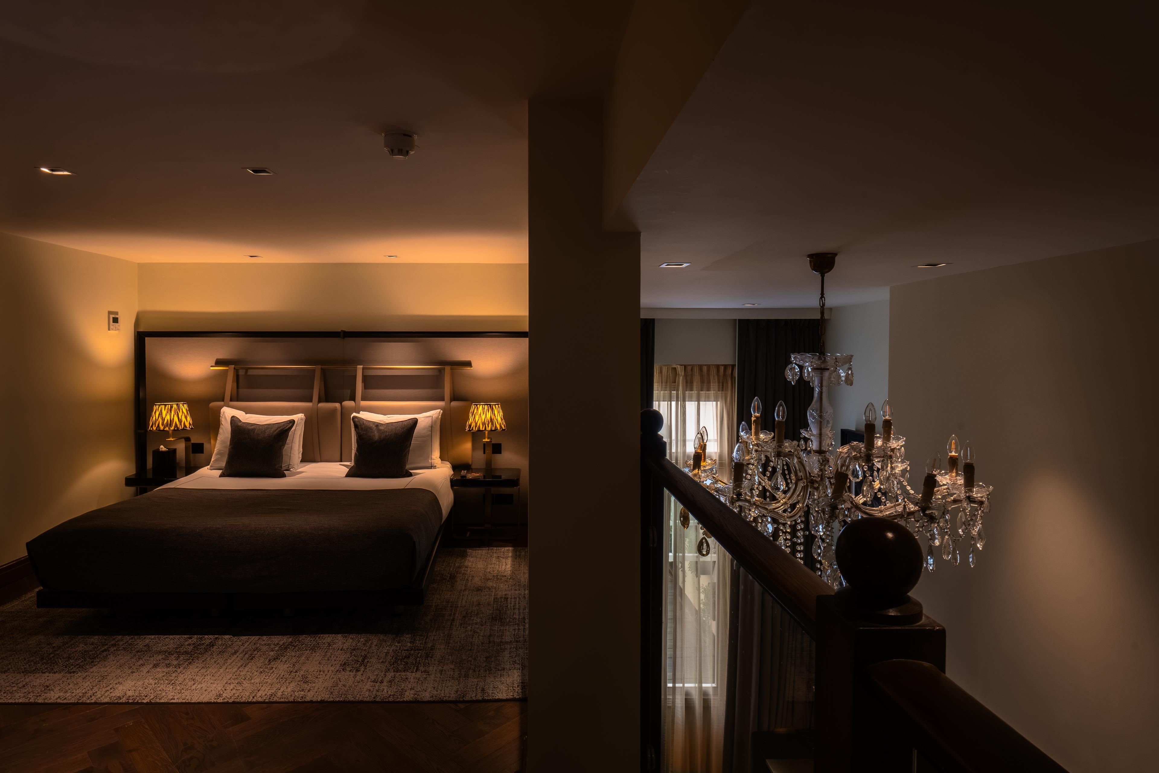 A hotel room with a large bed, bedside lamps, a chandelier, and a view of the outside through a window.