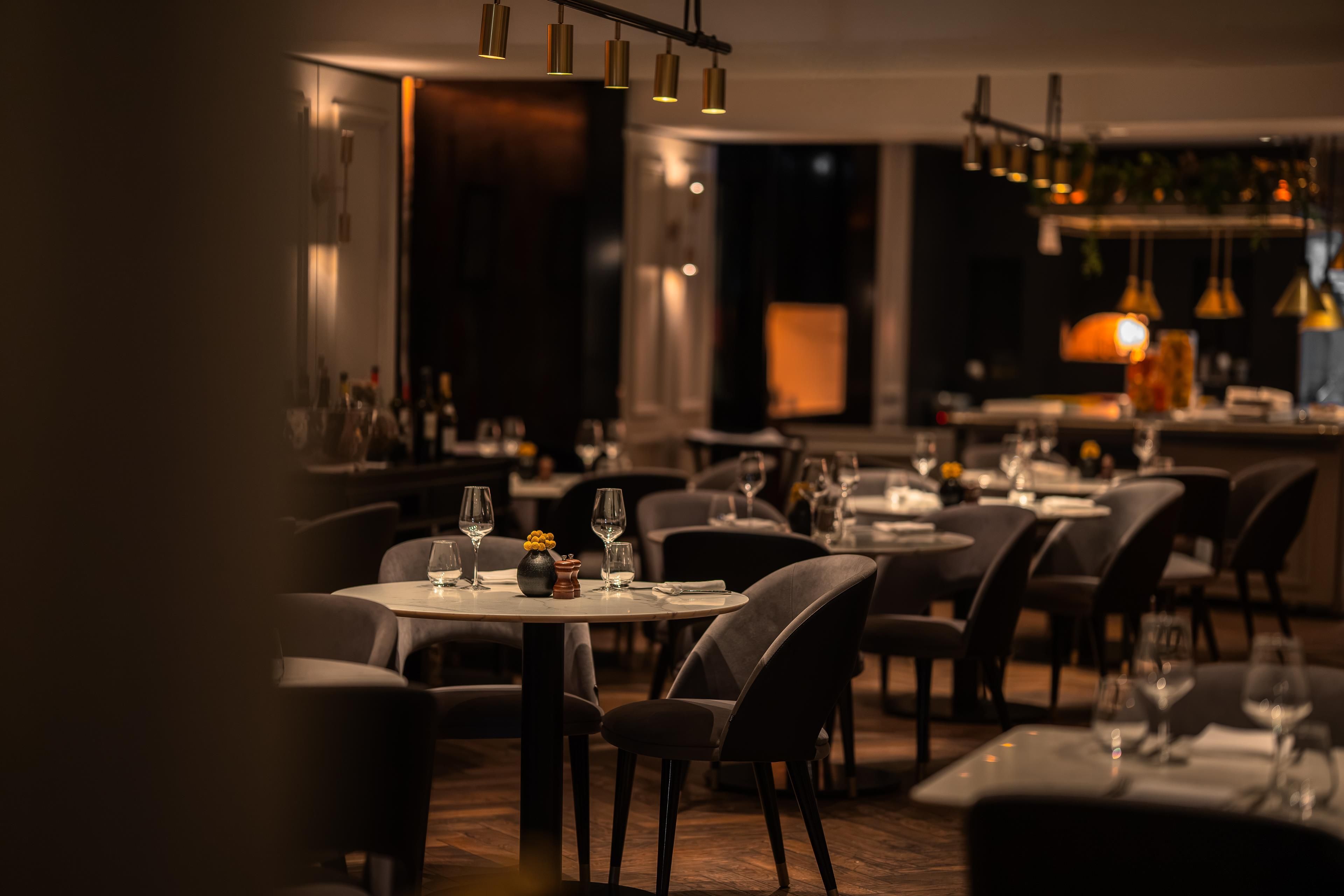 A dimly lit restaurant with round tables and chairs, a bar area, and pendant lighting.