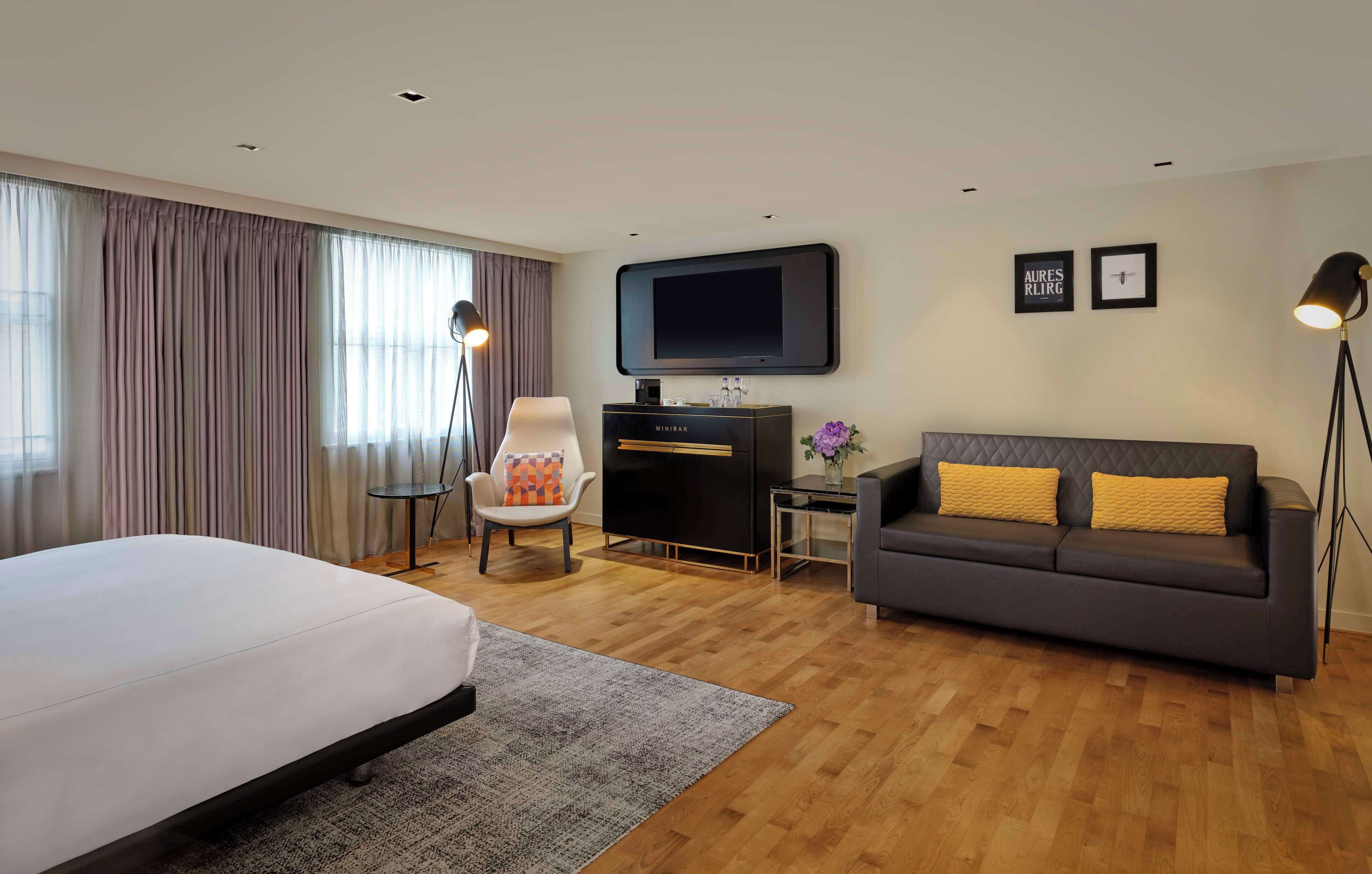 A hotel room with a bed, a sofa, a desk with a flat-screen TV, a chair, and a floor lamp.