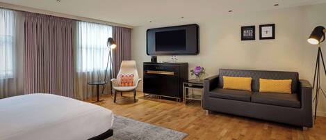 A hotel room with a bed, a sofa, a desk with a flat-screen TV, a chair, and a floor lamp.