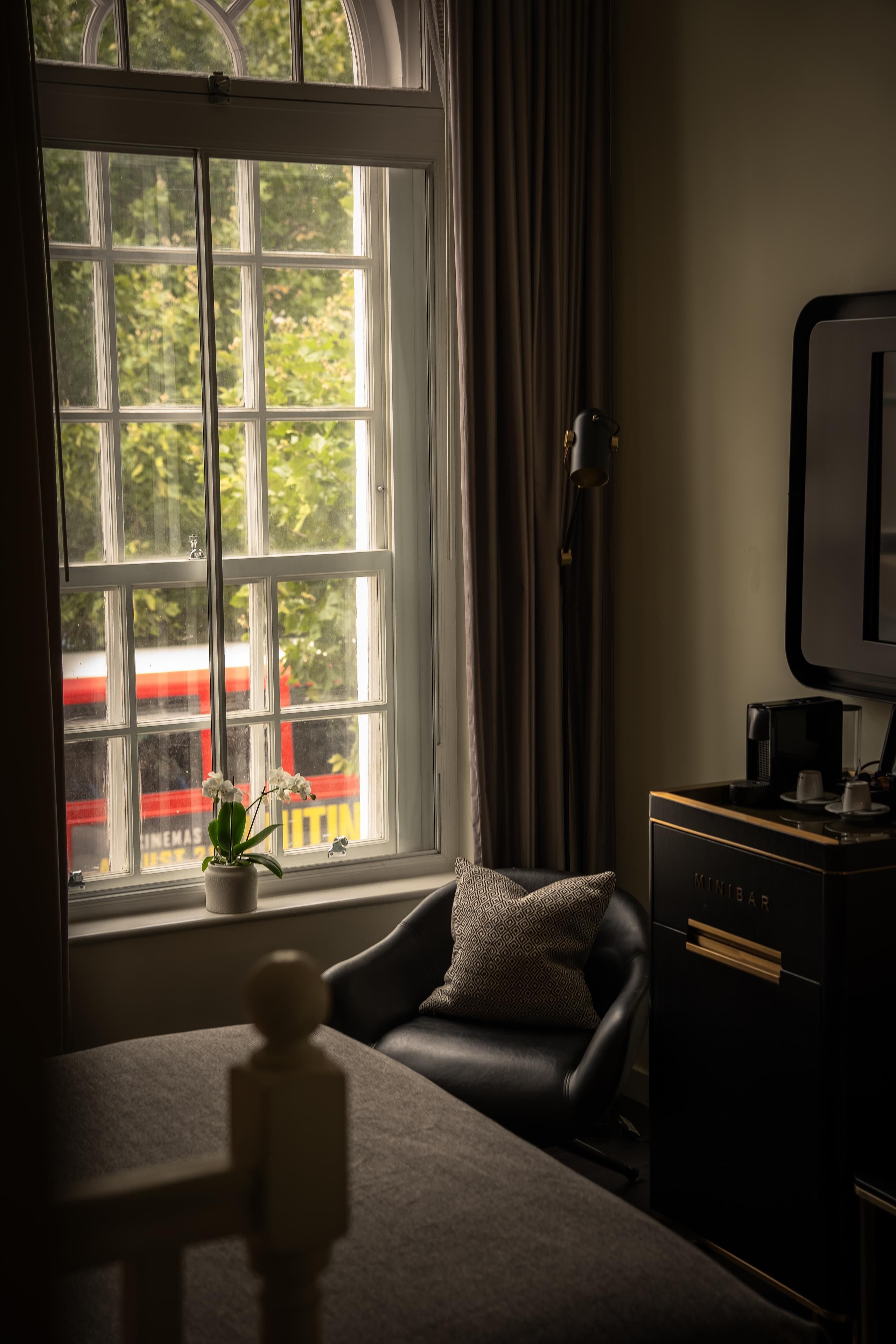 A room with a view of a red train through a window, a black leather chair, a small potted plant, and a black cabinet with a coffee maker.