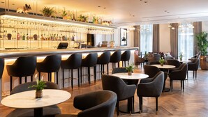 A modern bar area with a long counter, multiple bar stools, and a variety of bottles and glasses. There are several round tables with chairs, and potted plants are placed around the room. - Holmes Hotel London (London)