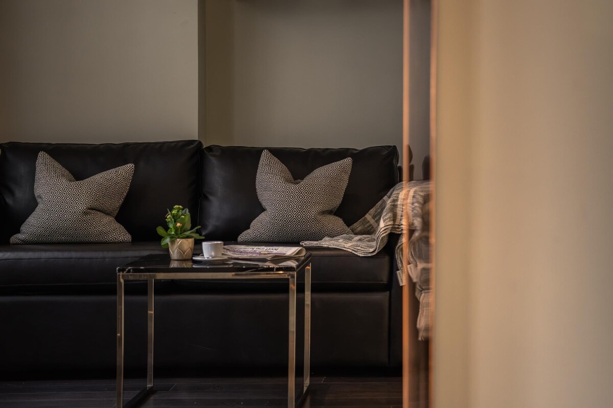 A modern living room with a black leather sofa, patterned cushions, a small table with a plant, and a framed artwork on the wall.