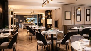 A modern dining area with round tables and chairs, a kitchen in the background, and framed artwork on the walls. - Holmes Hotel London (London)