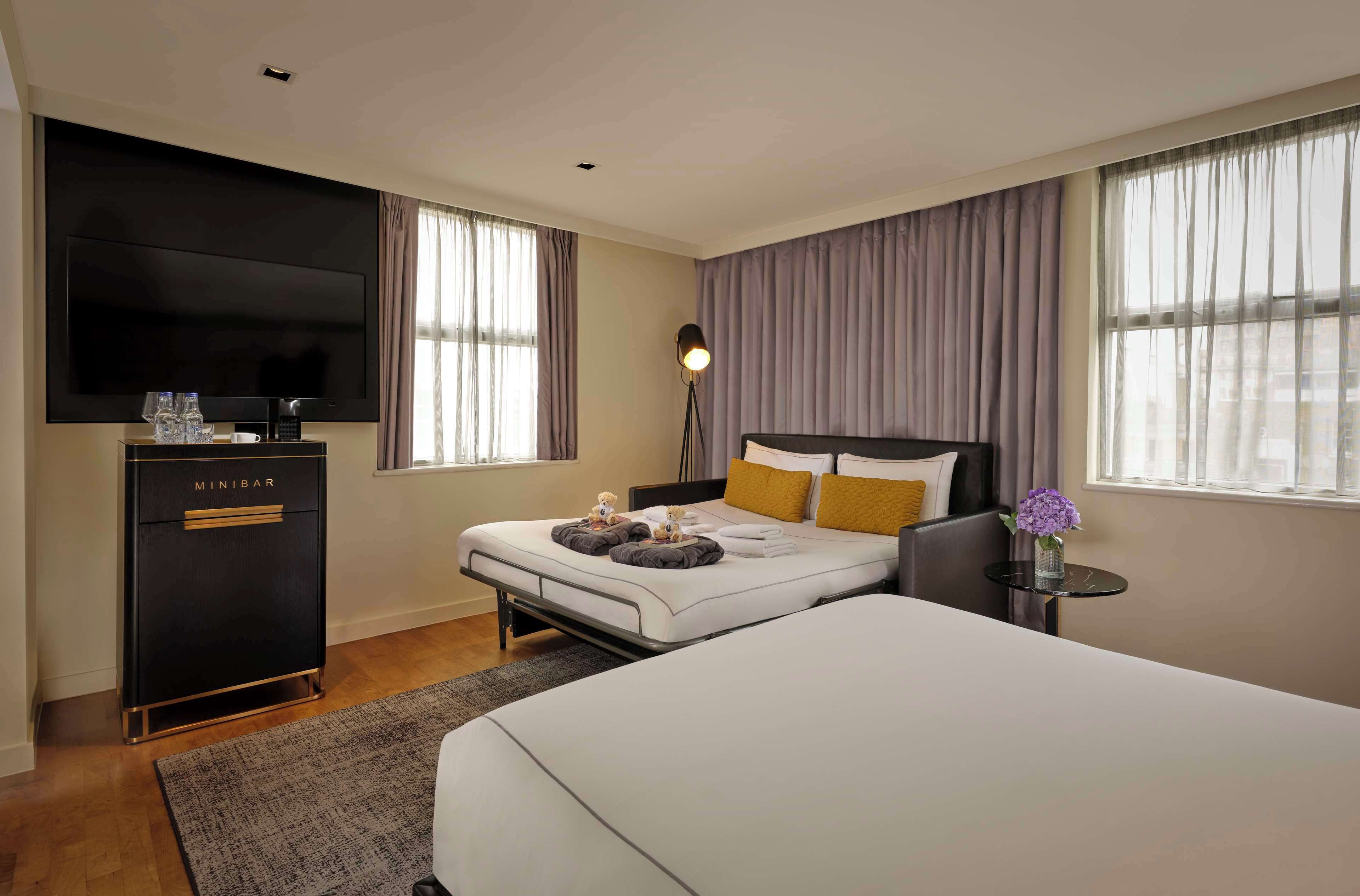 A hotel room with a bed, a TV, a minibar, and a bedside table with a lamp.