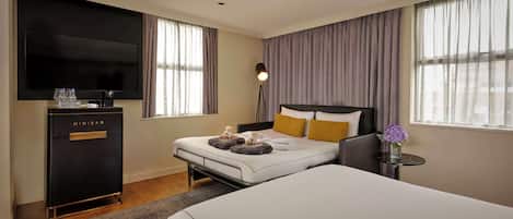 A hotel room with a bed, a TV, a minibar, and a bedside table with a lamp.
