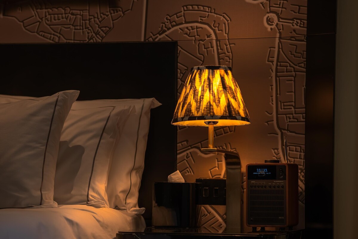 A hotel room with a bed, a nightstand, a lamp, and a wall with a map design.