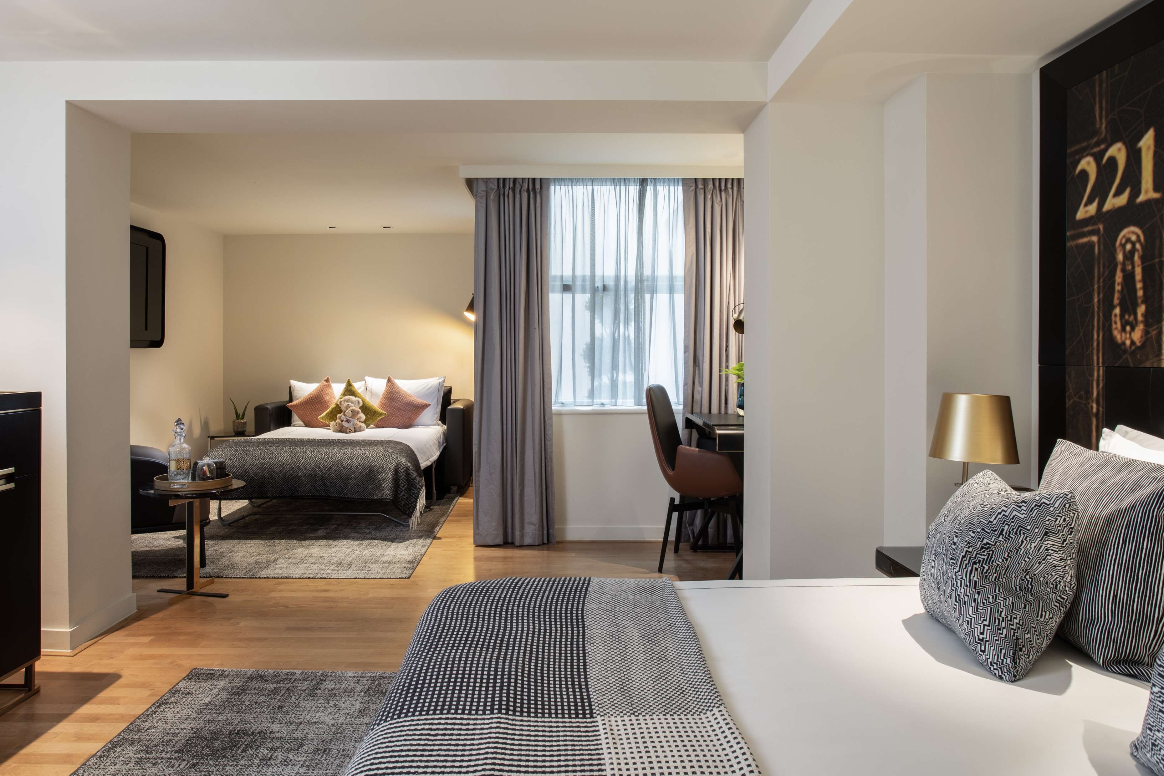 A modern hotel room with a large bed, a desk, and a chair. There is a window with curtains, a lamp, and a wall-mounted TV.