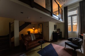 A spacious living room with a leather sofa, a coffee table, and a bookshelf. A staircase leads to an upper level, and a large window provides natural light. - Holmes Hotel London (London)