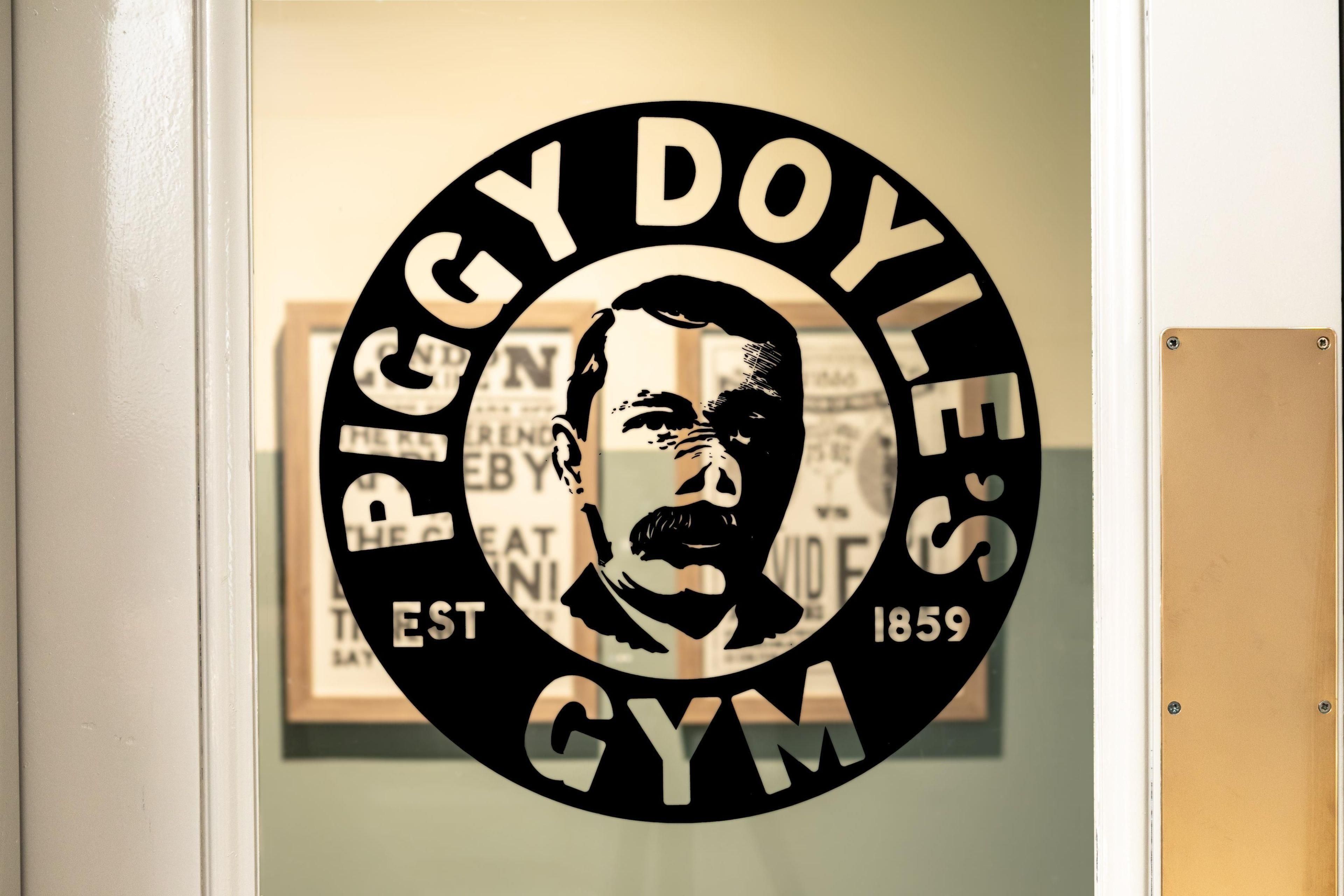Ein rundes Schild mit der Silhouette eines Mannes und dem Text 'Piggy Doyle's Gym' und 'GEGR. 1859'.