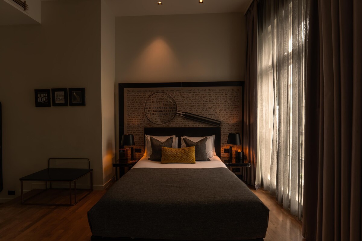 A modern bedroom with a large bed, bedside lamps, a hanging light fixture, and a view of the outdoors through sheer curtains.
