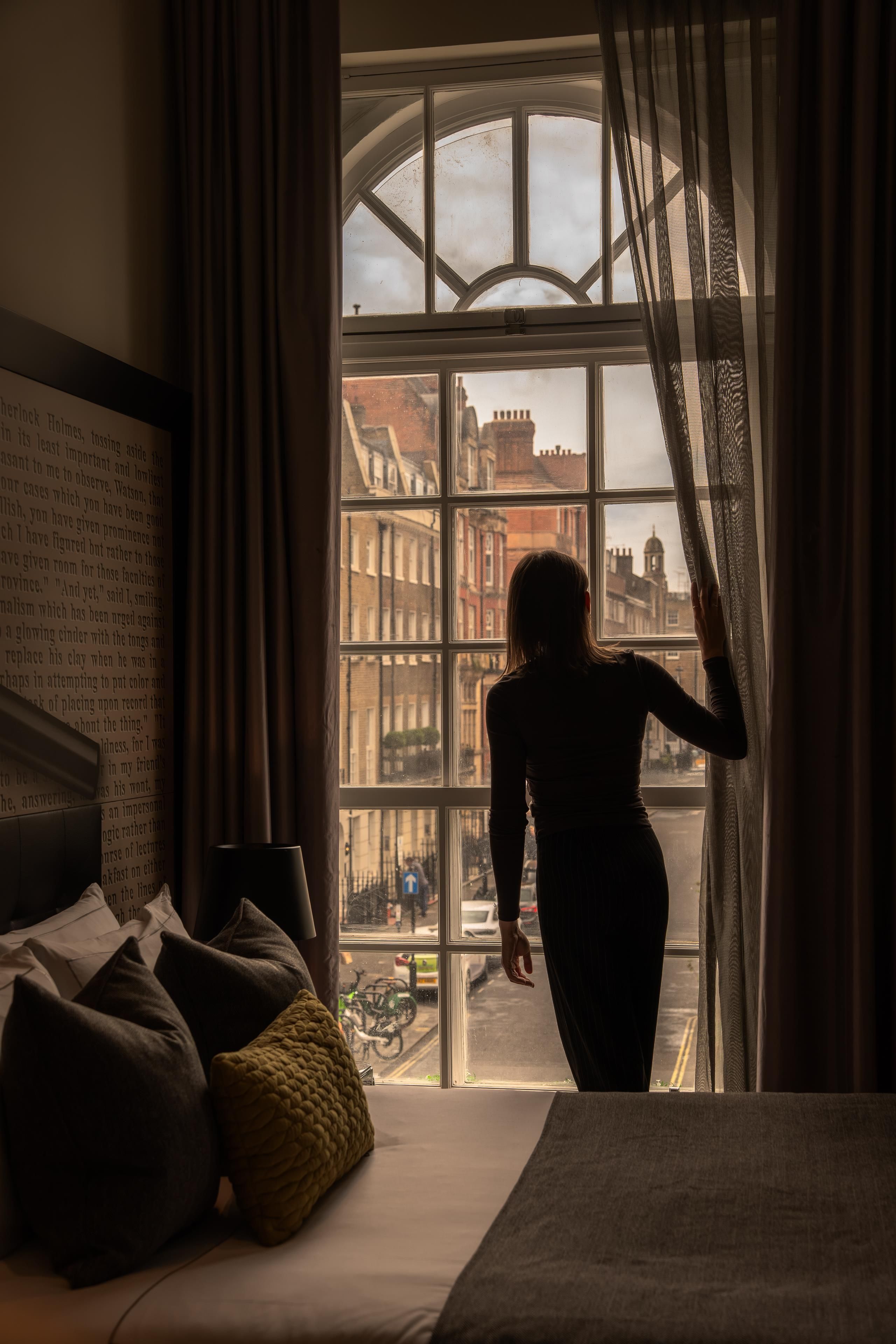 Ein Zimmer mit großem Fenster, einem Bett und einer Person, die am Fenster steht.