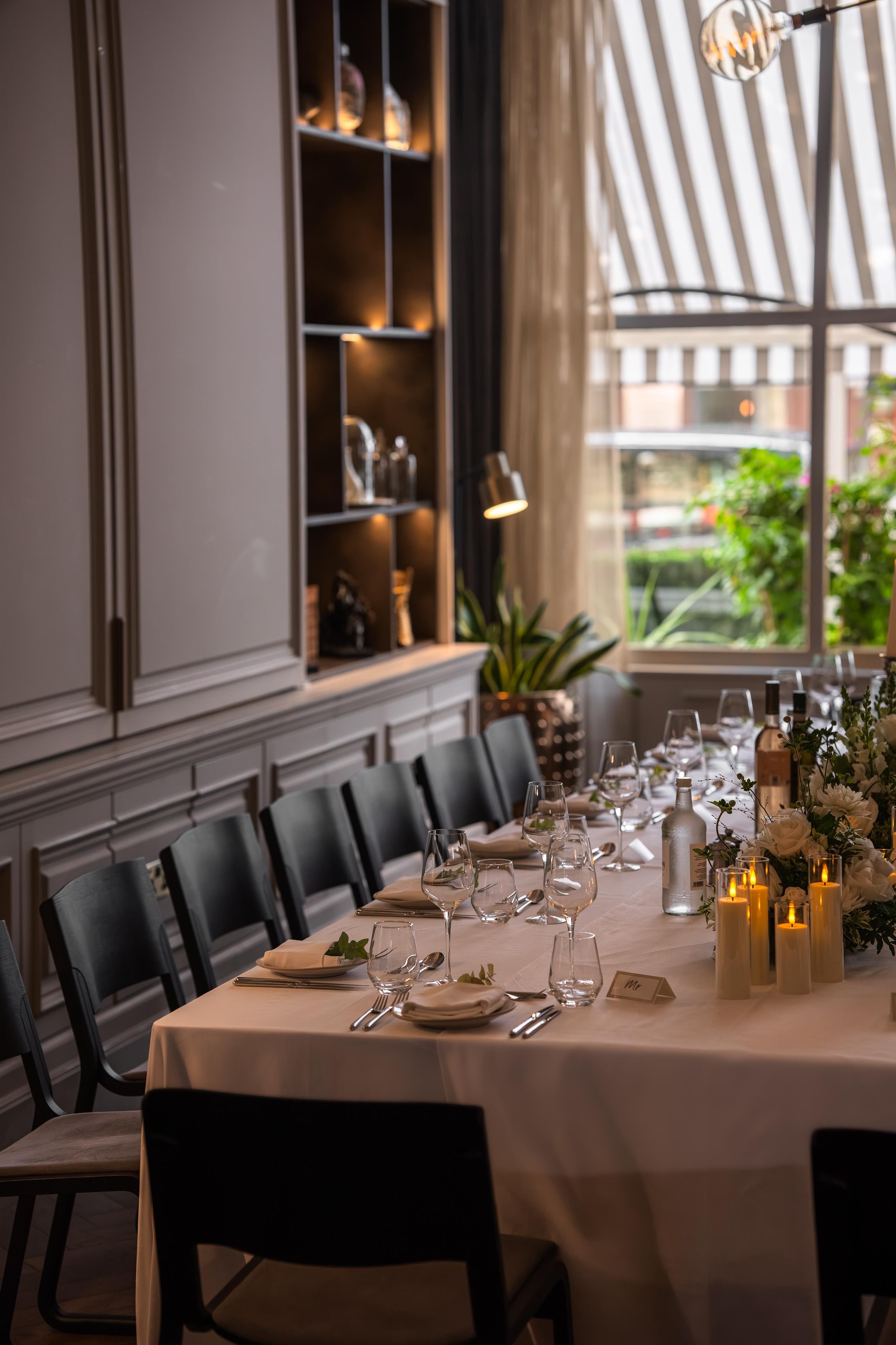 A dining table set for a meal with chairs, candles, and a view of greenery outside.