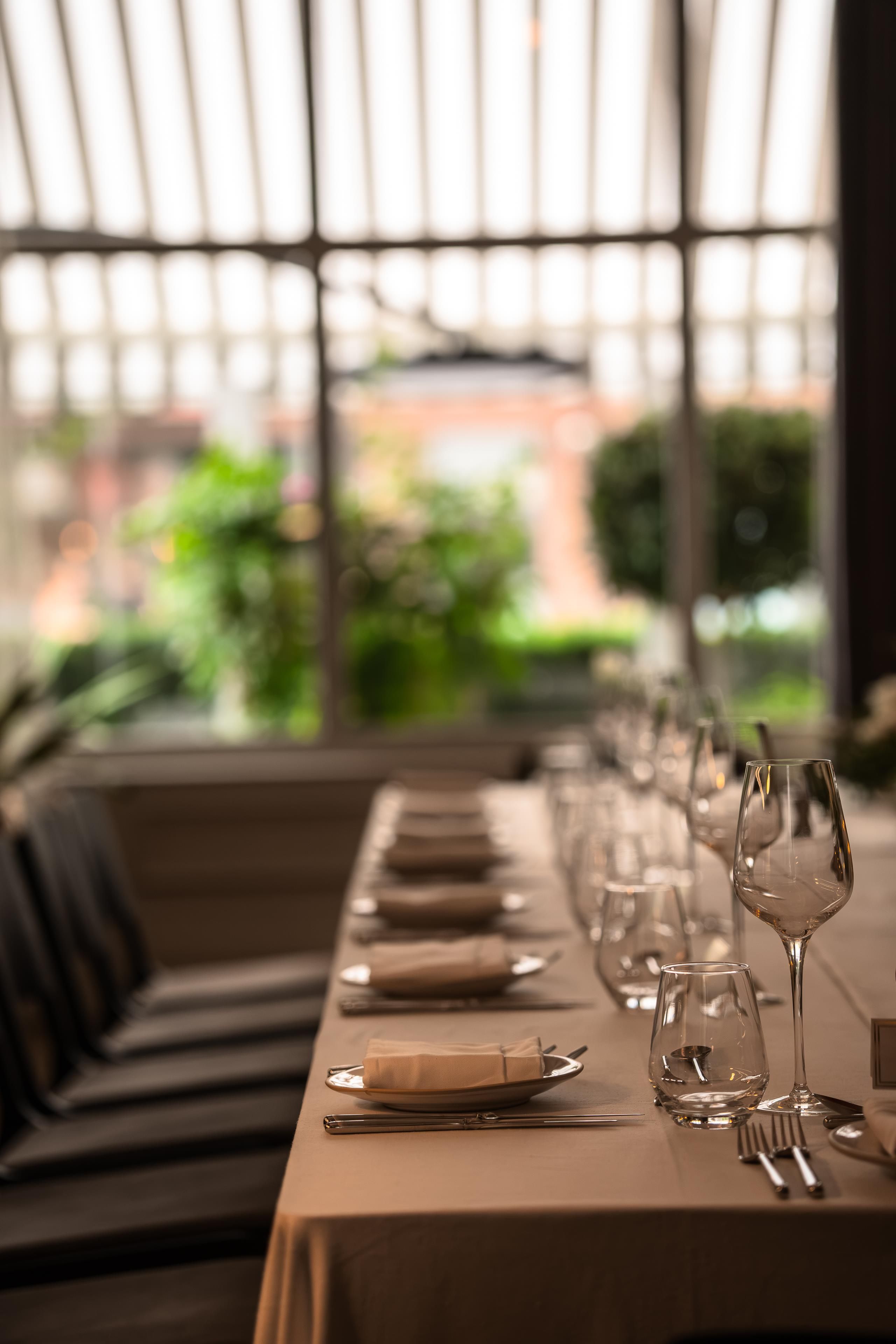 A dining table set with plates, cutlery, and wine glasses, with a view of greenery and a building through a window.