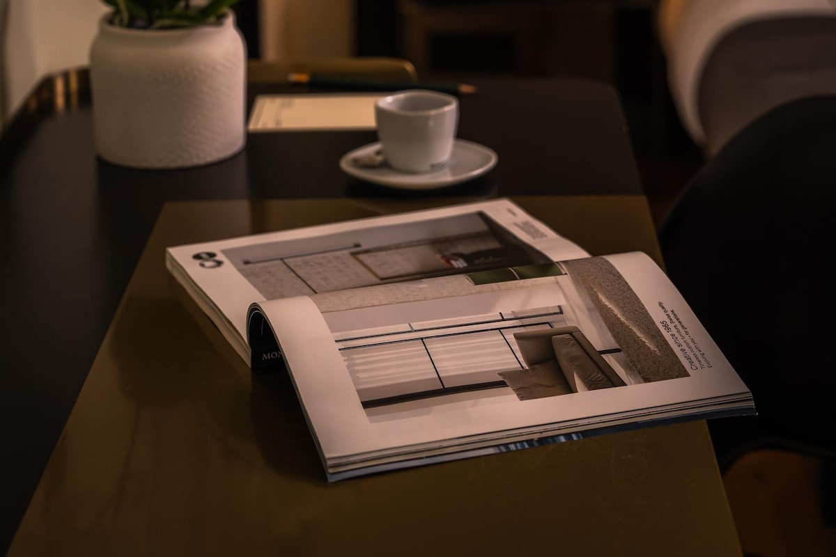 A hotel room with a bed, a nightstand with a lamp, a table with a cup and saucer, and a potted plant.