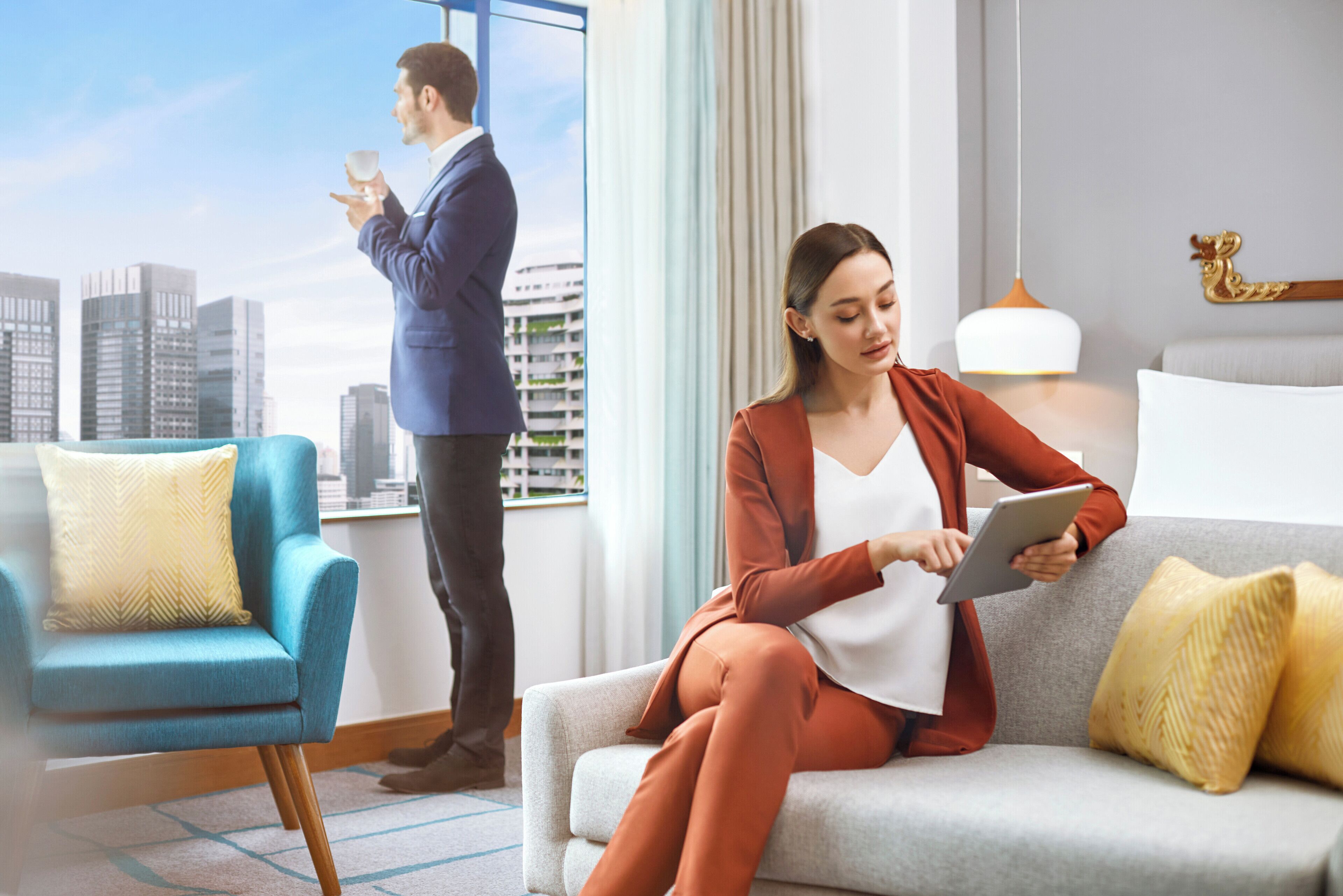 A modern hotel room with a city view, a sofa, a chair, and a tablet.