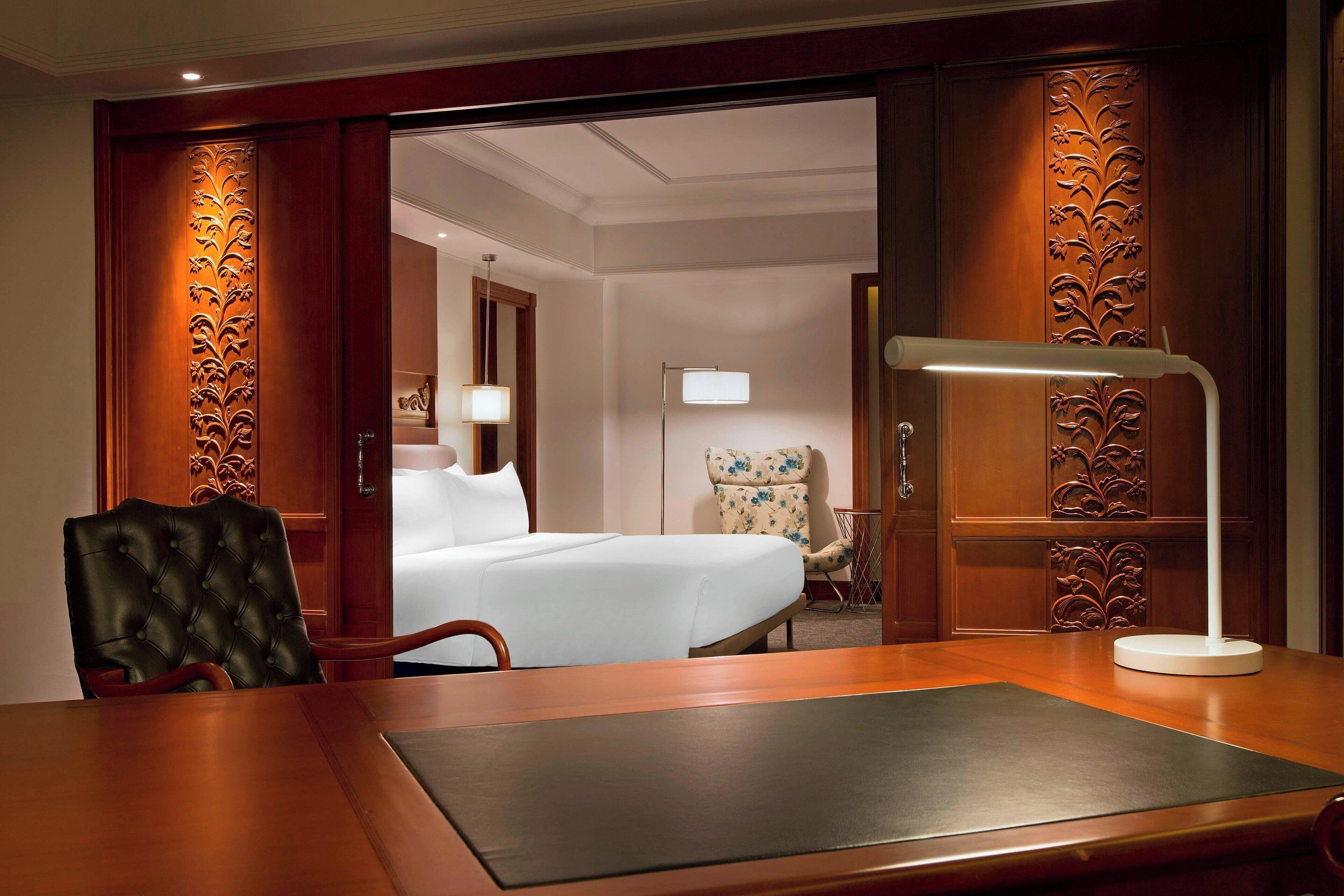 A hotel room with a bed, a desk, a chair, and a lamp.