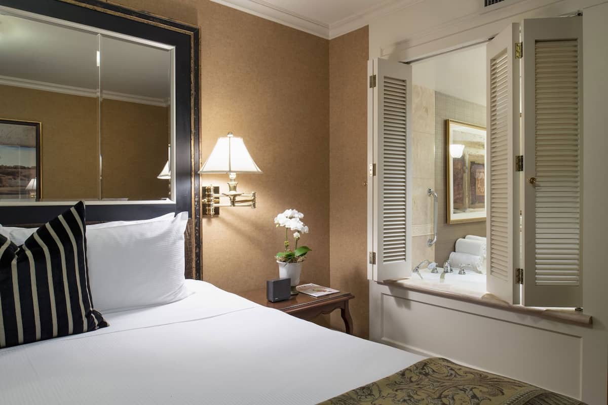 Habitación de hotel con cama, una lámpara de noche, una mesita con una planta, una ventana con persianas y un espejo.