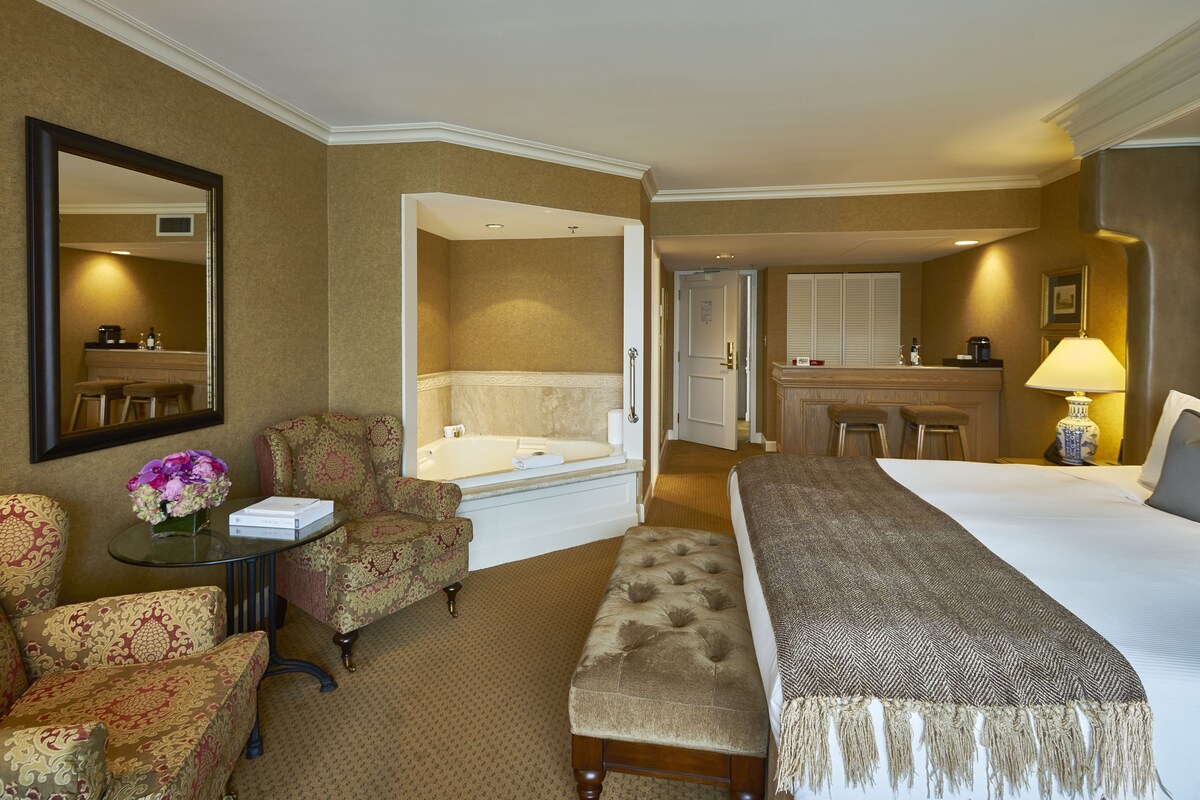 A hotel room with a bed, a sitting area with a chair and a small table, a bathroom with a bathtub, and a kitchenette.