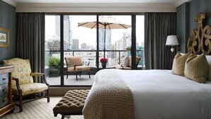 A hotel room with a large bed, a chair, a small table with a potted plant, and a view of the cityscape through large windows. - Wedgewood Hotel & Spa (Vancouver)
