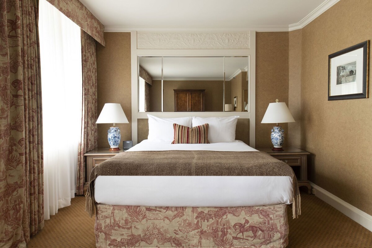 A hotel room with a large bed, two bedside tables with lamps, a mirror above the bed, and a patterned carpet.