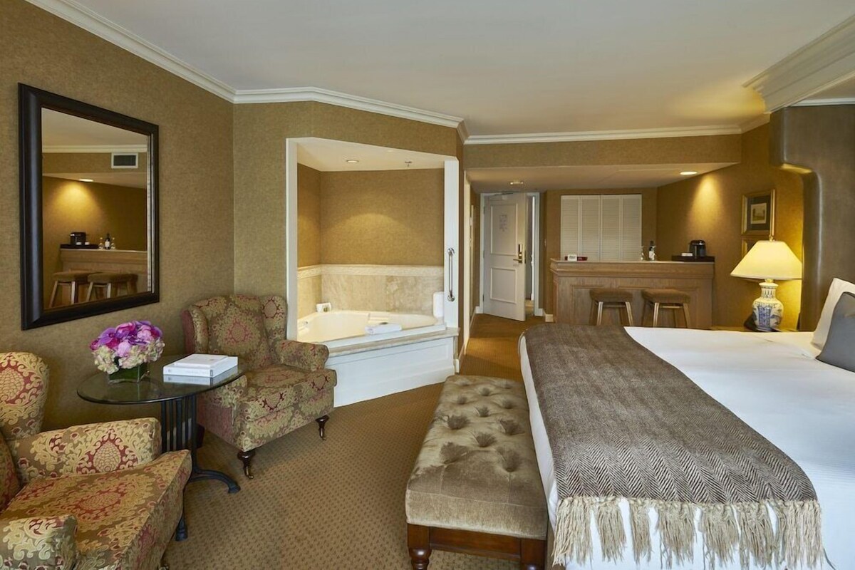 A hotel room with a bed, a sitting area with a chair and a small table, a bathroom with a bathtub, and a kitchenette.