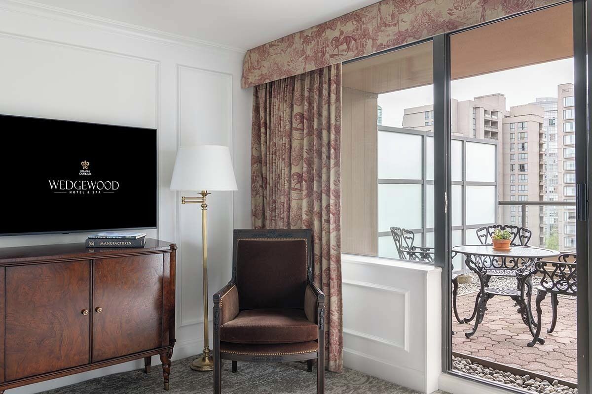 A hotel room with a television, a chair, a lamp, a window with curtains, and a balcony with a table and chairs.