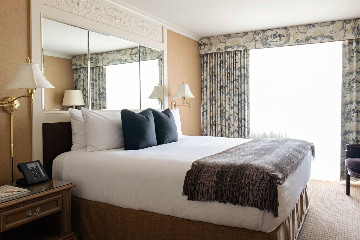 A hotel room with a large bed, two bedside lamps, a nightstand with a phone, a chair, and a large mirror.