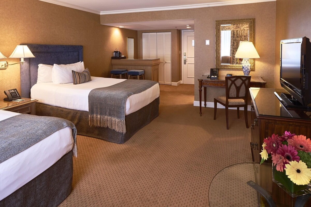 A hotel room with two beds, a desk, a chair, a television, and a bathroom visible in the background.