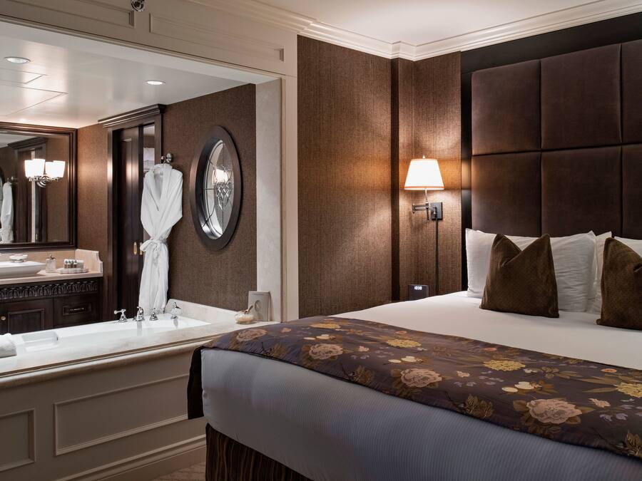 A hotel room with a large bed, a nightstand, a mirror, a bathroom with a sink and vanity, and a wall clock.