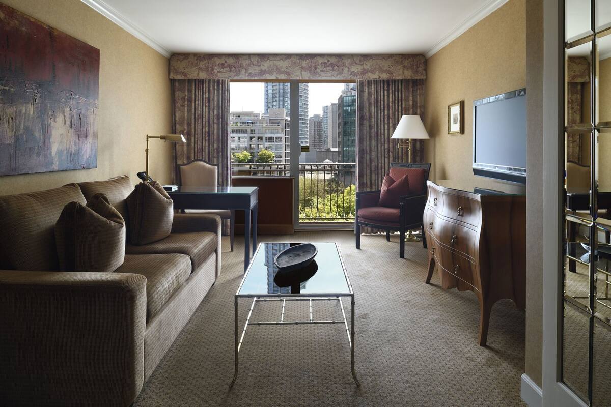 Habitación de hotel con sofá, mesa de centro, sillón, televisión, cómoda y balcón con vista a la ciudad.