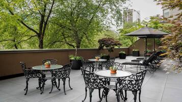 Un patio al aire libre con mesas y sillas redondas, rodeado de plantas en macetas y árboles.