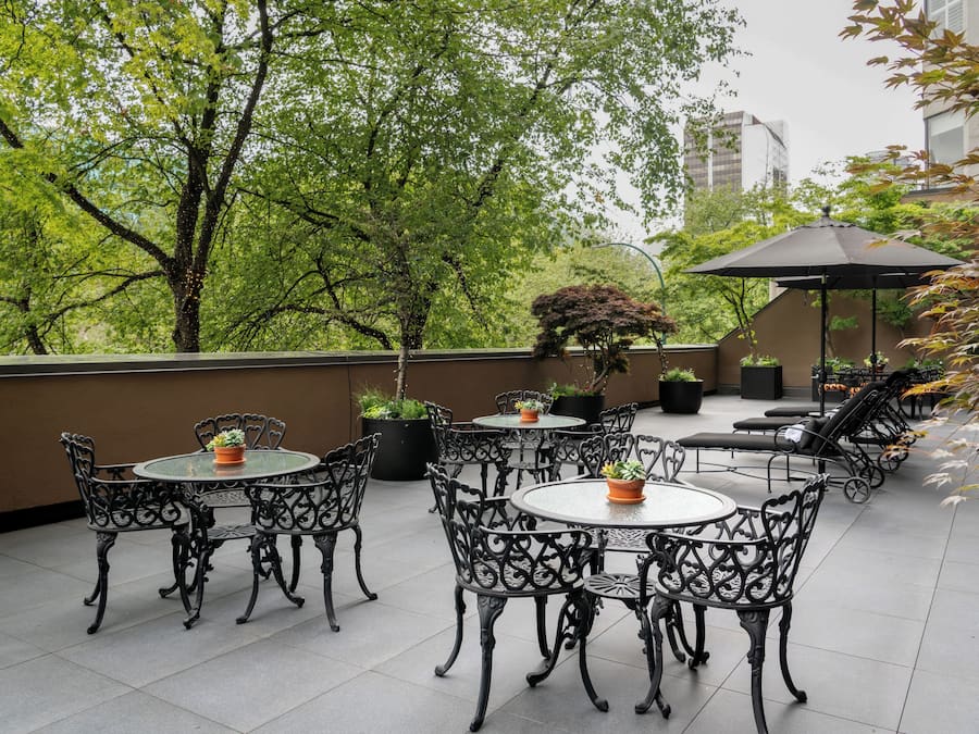 Un patio al aire libre con mesas y sillas redondas, rodeado de plantas en macetas y árboles.