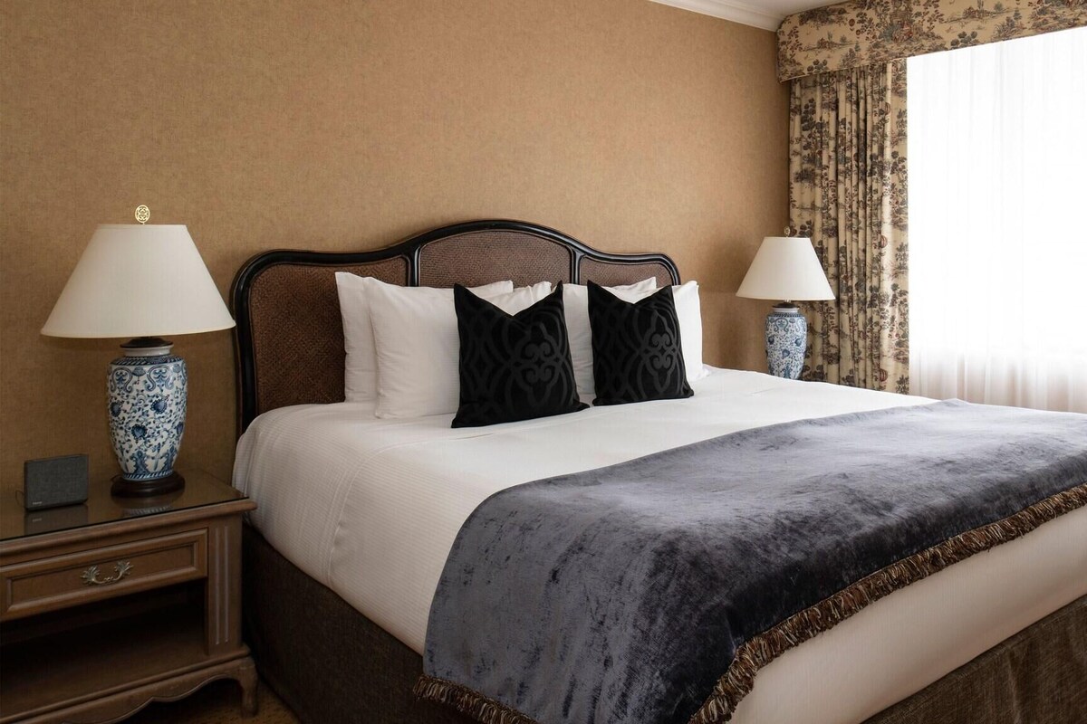 A hotel room with a bed, two bedside tables, lamps, a nightstand, and a window with curtains.