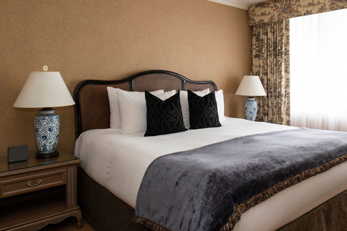 A hotel room with a large bed, two bedside tables, a nightstand, a decorative lamp, and a window with curtains.
