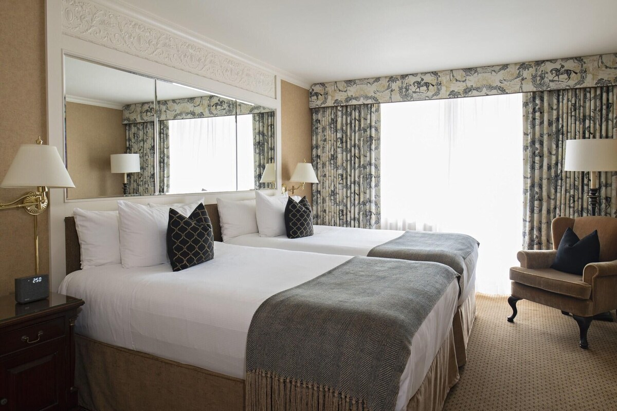 A hotel room with two beds, a large mirror, a chair, and a nightstand with a lamp.