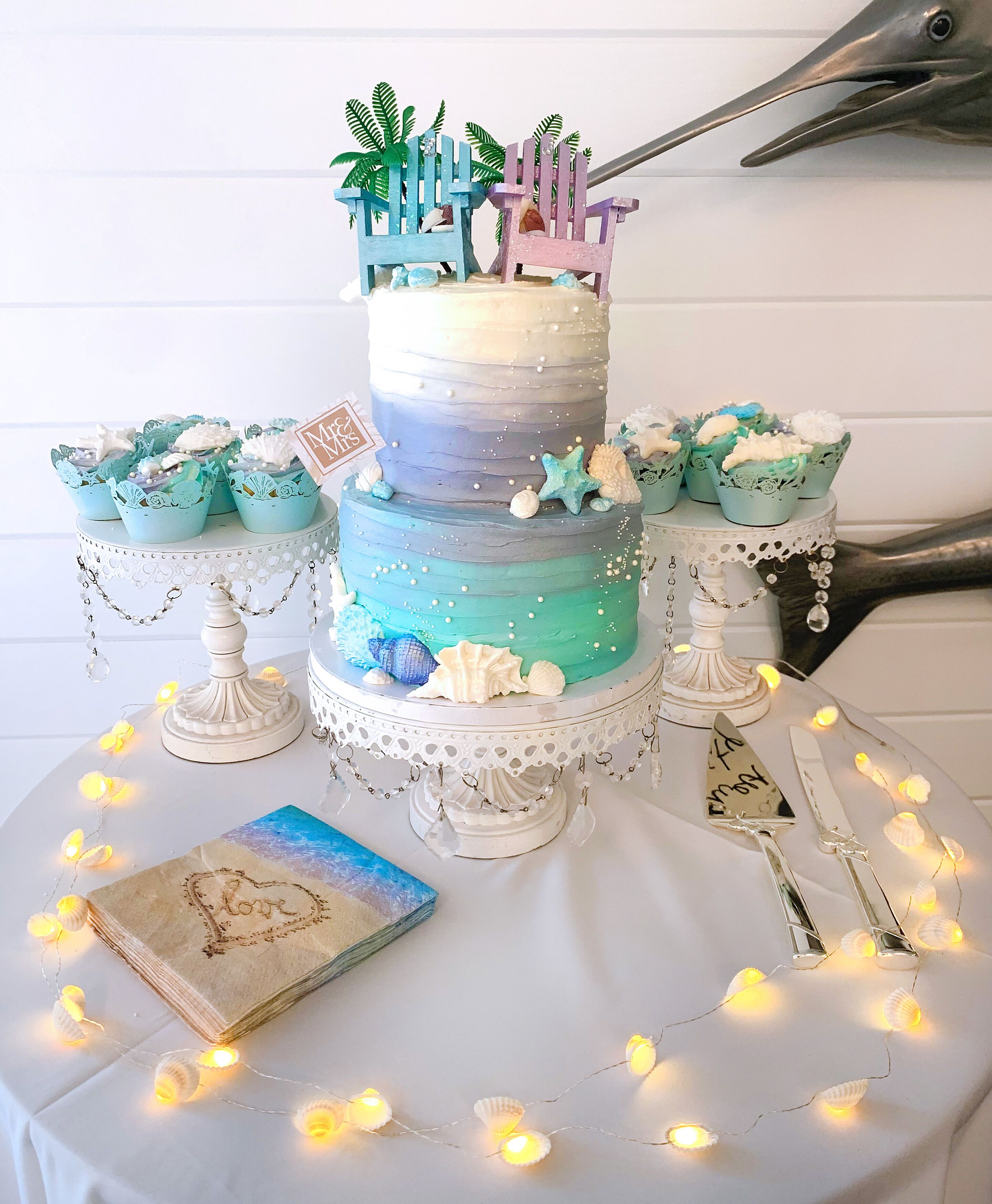 A multi-tiered cake with a tropical theme, surrounded by cupcakes and decorative elements on a table.