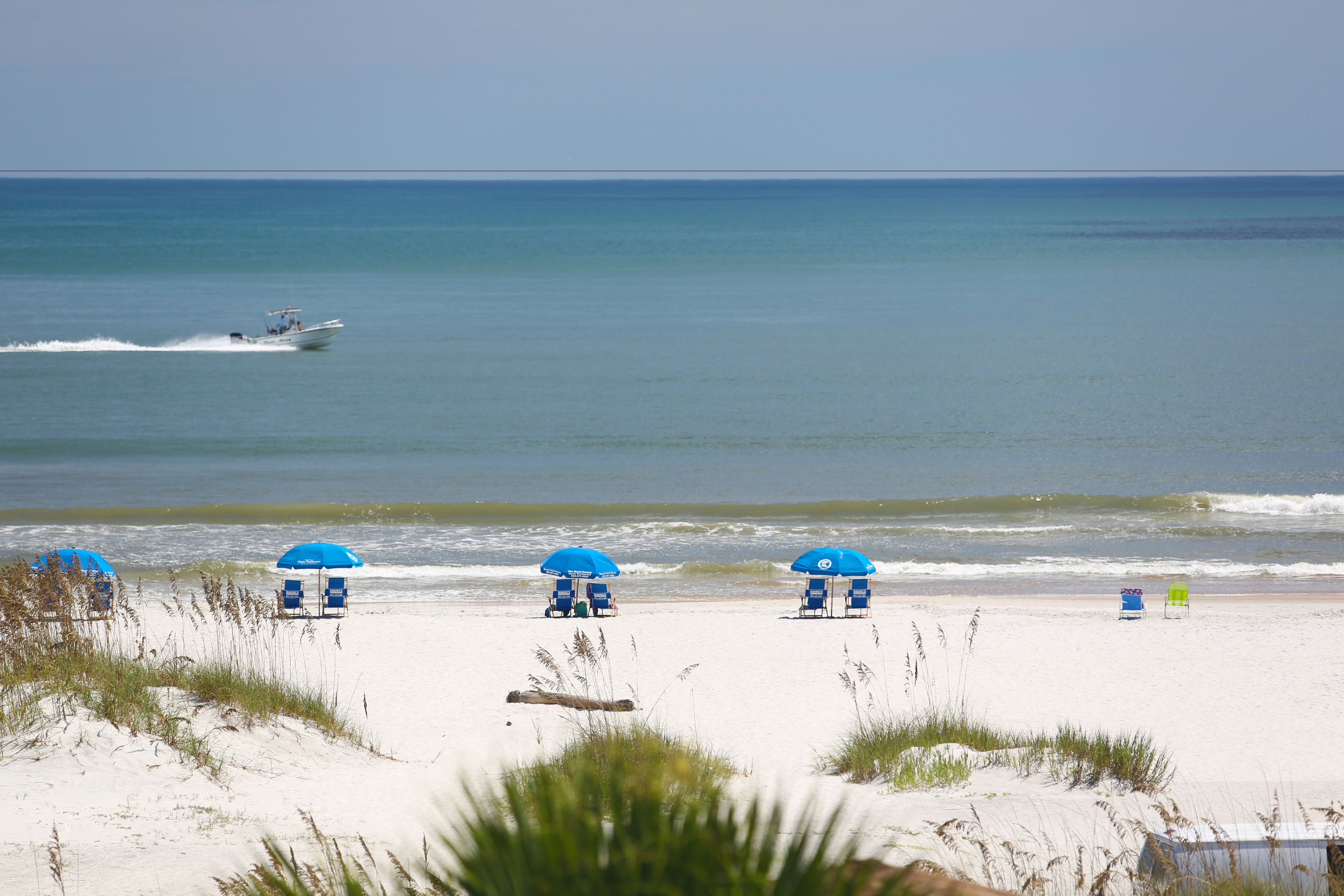 A beach with white sand, blue umbrellas, and a boat in the water.