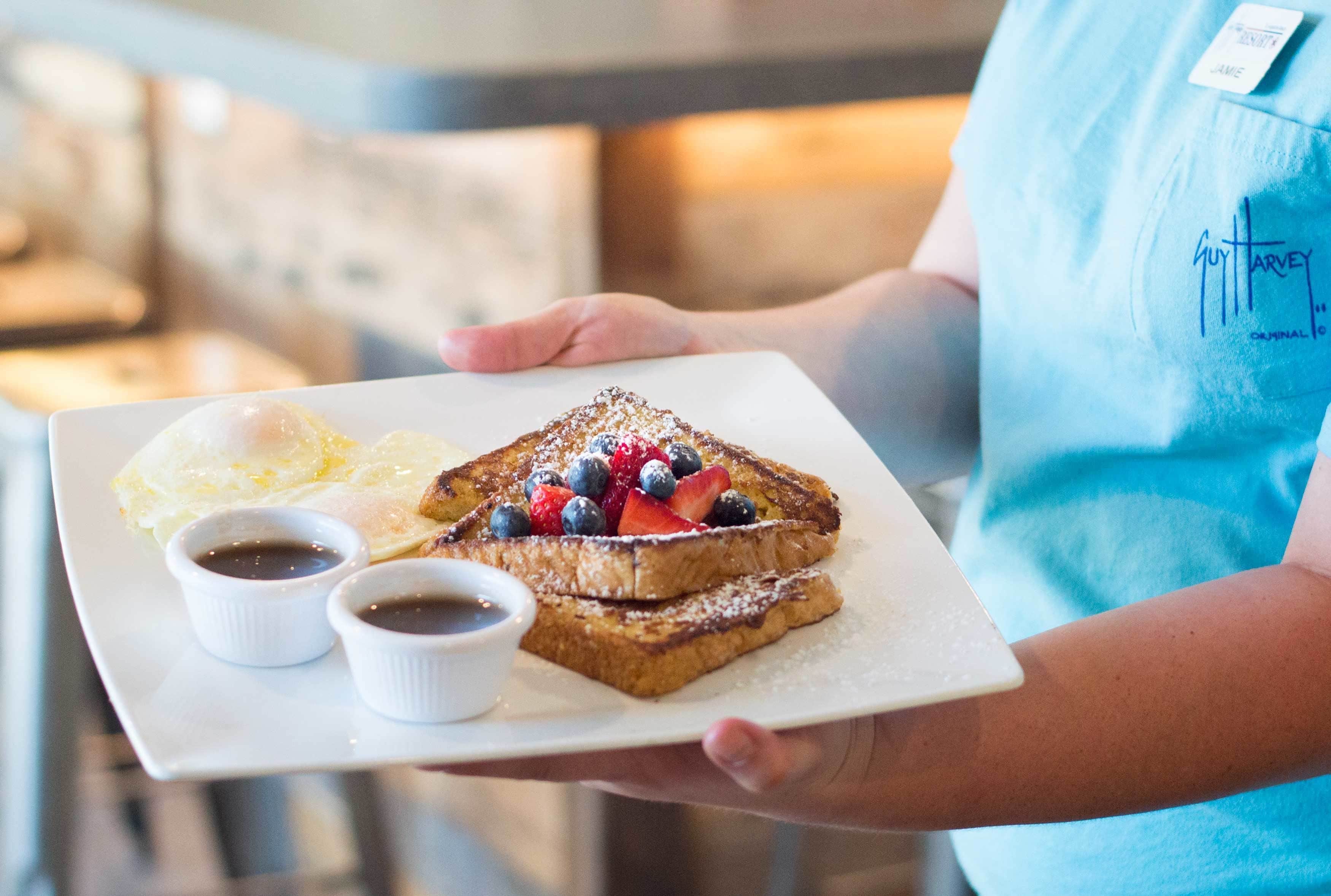 A person holding a plate with French toast, strawberries, blueberries, and two small cups of syrup.