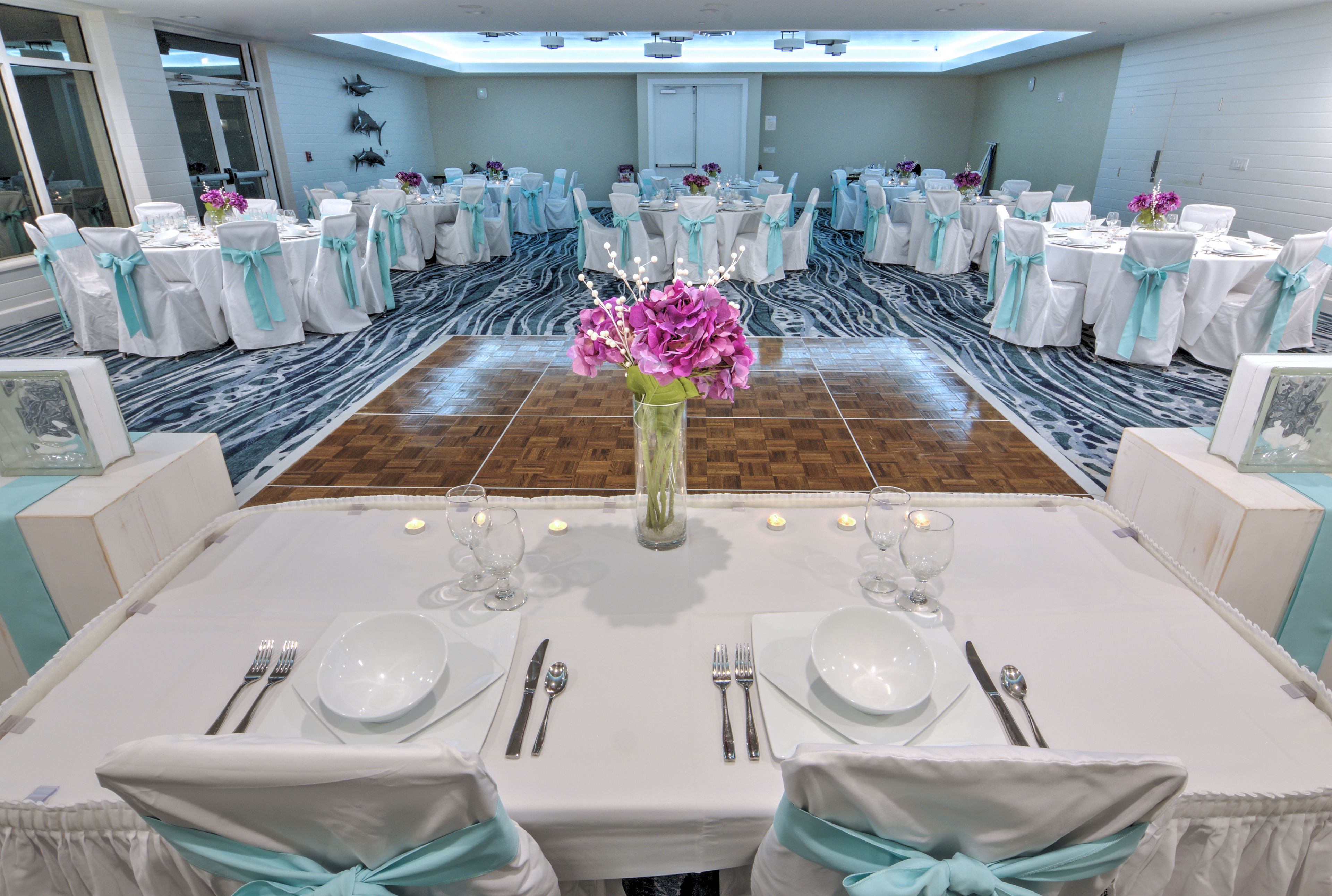 Un salón de banquetes con mesas redondas, manteles blancos y cintas teal.