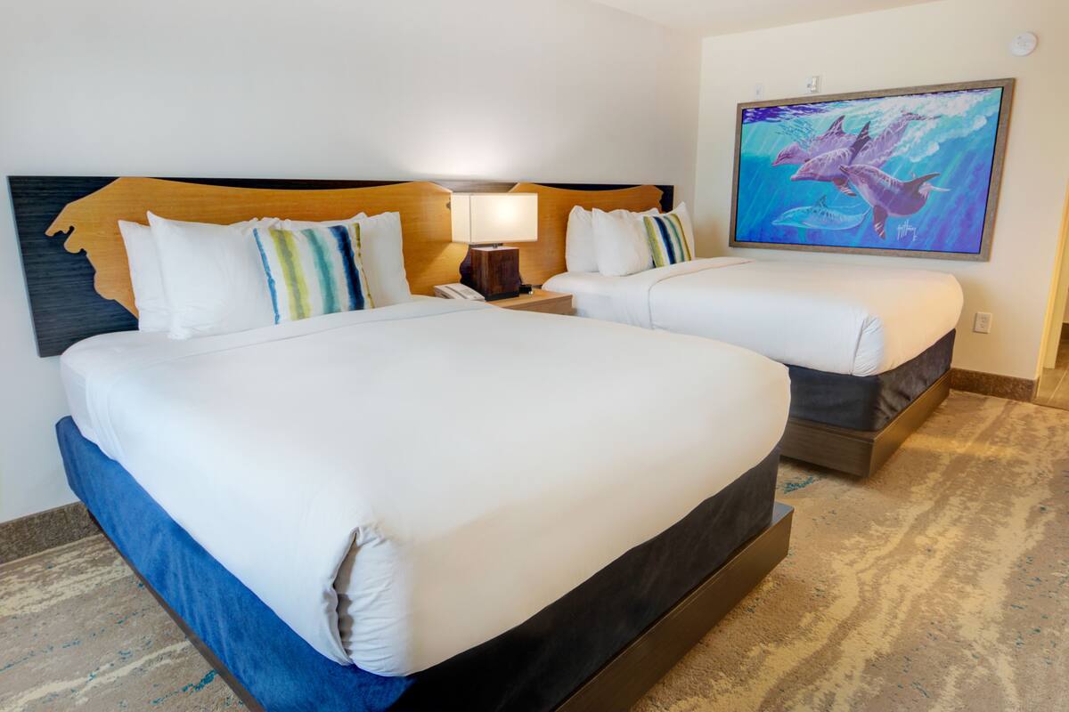 Een hotelkamer met twee bedden, een houten hoofdbord, een nachtkastje met een lamp, en een schilderij van dolfijnen aan de muur.