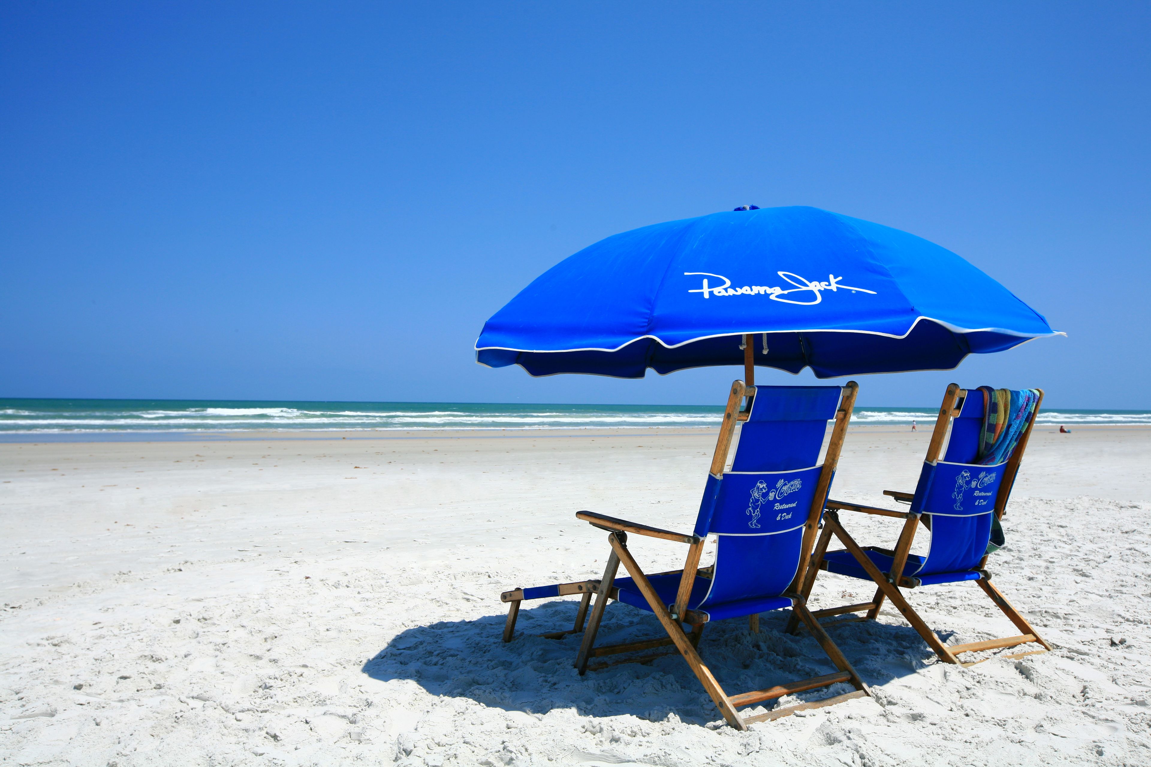 Una silla de playa y una sombrilla con la leyenda "Pavement Jack", sobre una playa arenosa con el océano al fondo.