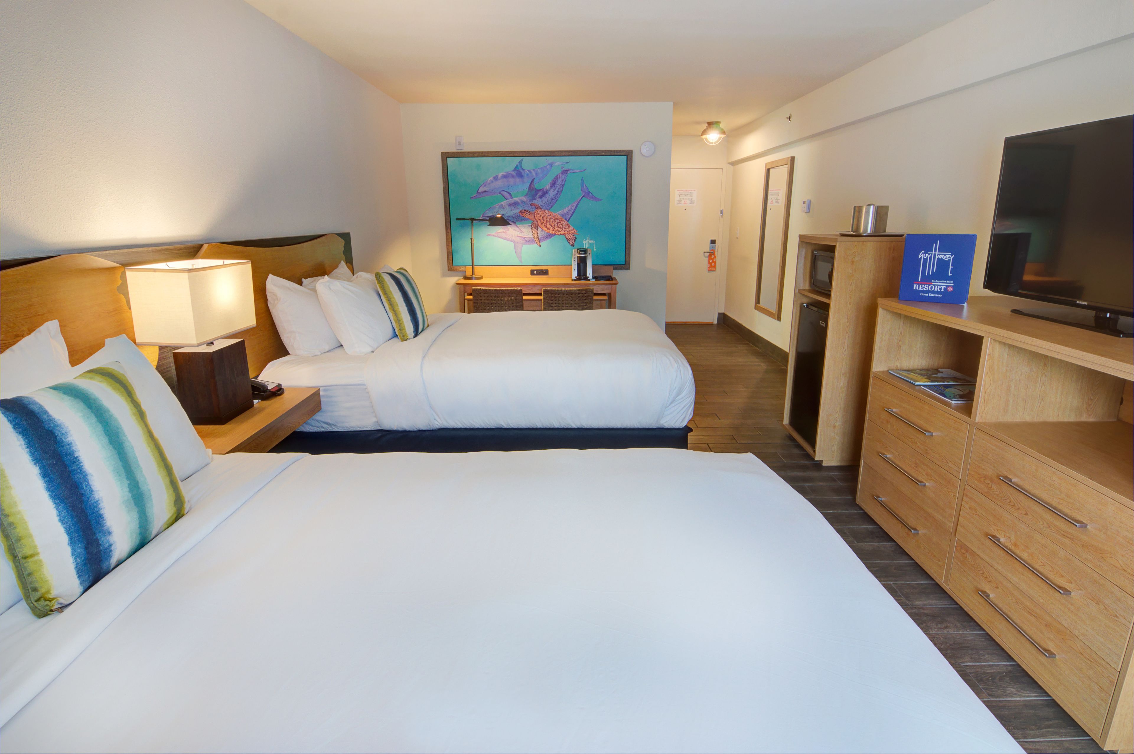 Ein Hotelzimmer mit zwei Betten, einem Schreibtisch mit Lampe, einem Fernseher und einem Bild von Delfinen an der Wand.