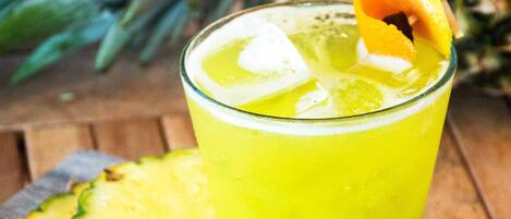 A glass of yellow beverage with ice cubes and a slice of pineapple on the rim, placed on a wooden board with a sprig of rosemary.