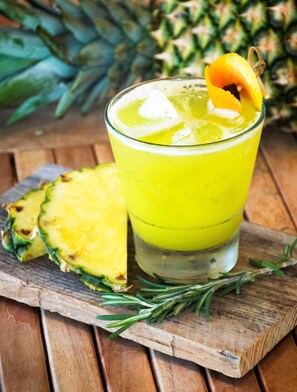 Un verre contenant une boisson jaune, avec des glaçons et une tranche d’ananas sur le bord, posé sur une planche en bois, agrémenté d’une branche de romarin.