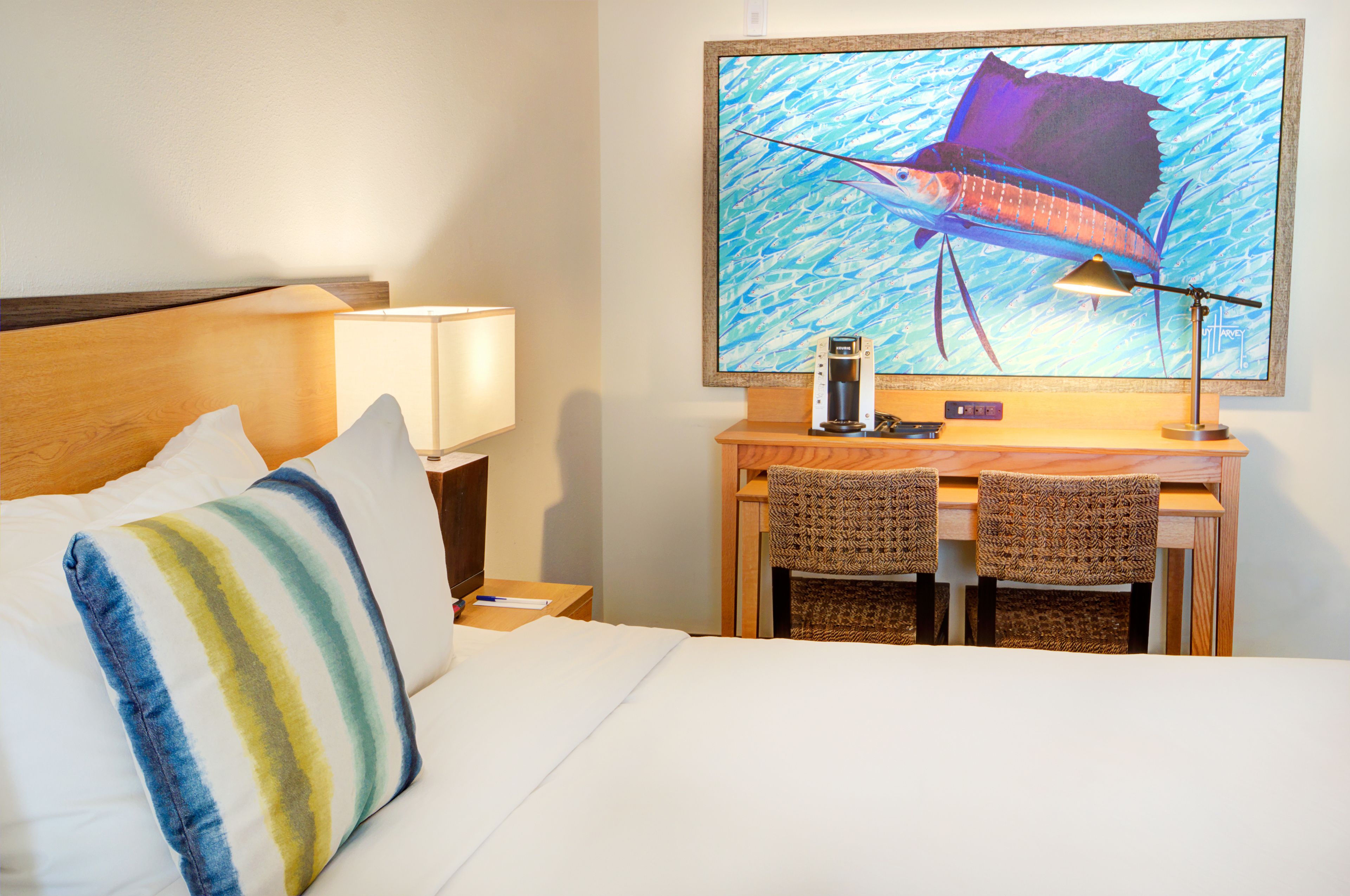 A hotel room with a bed, a desk with two chairs, a lamp, and a large framed picture of a fish on the wall.