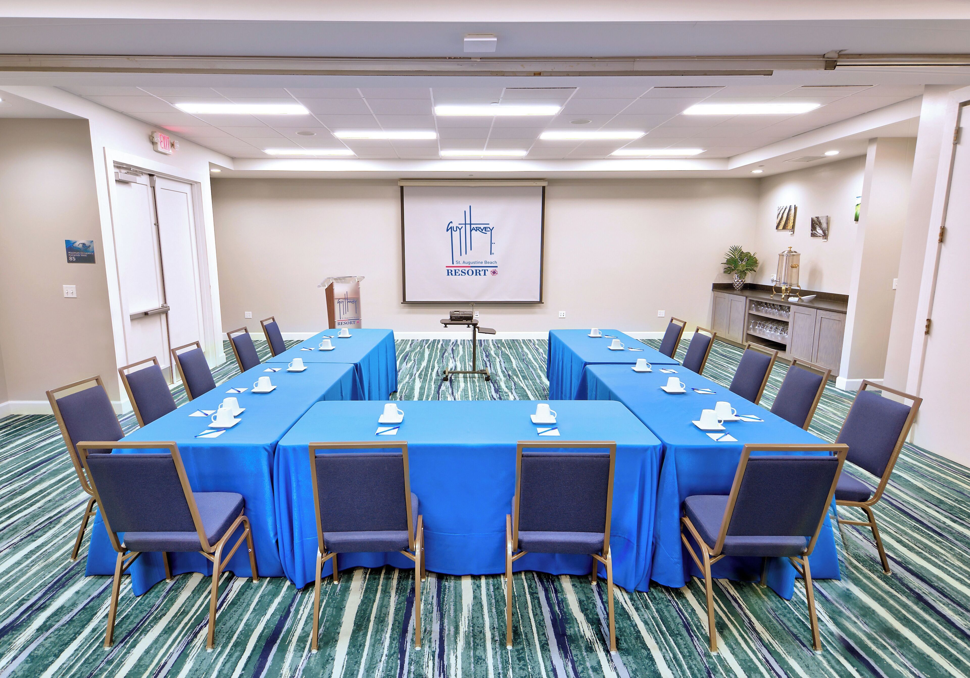 A conference room with a long table, chairs, a projector screen, and a small kitchenette area.