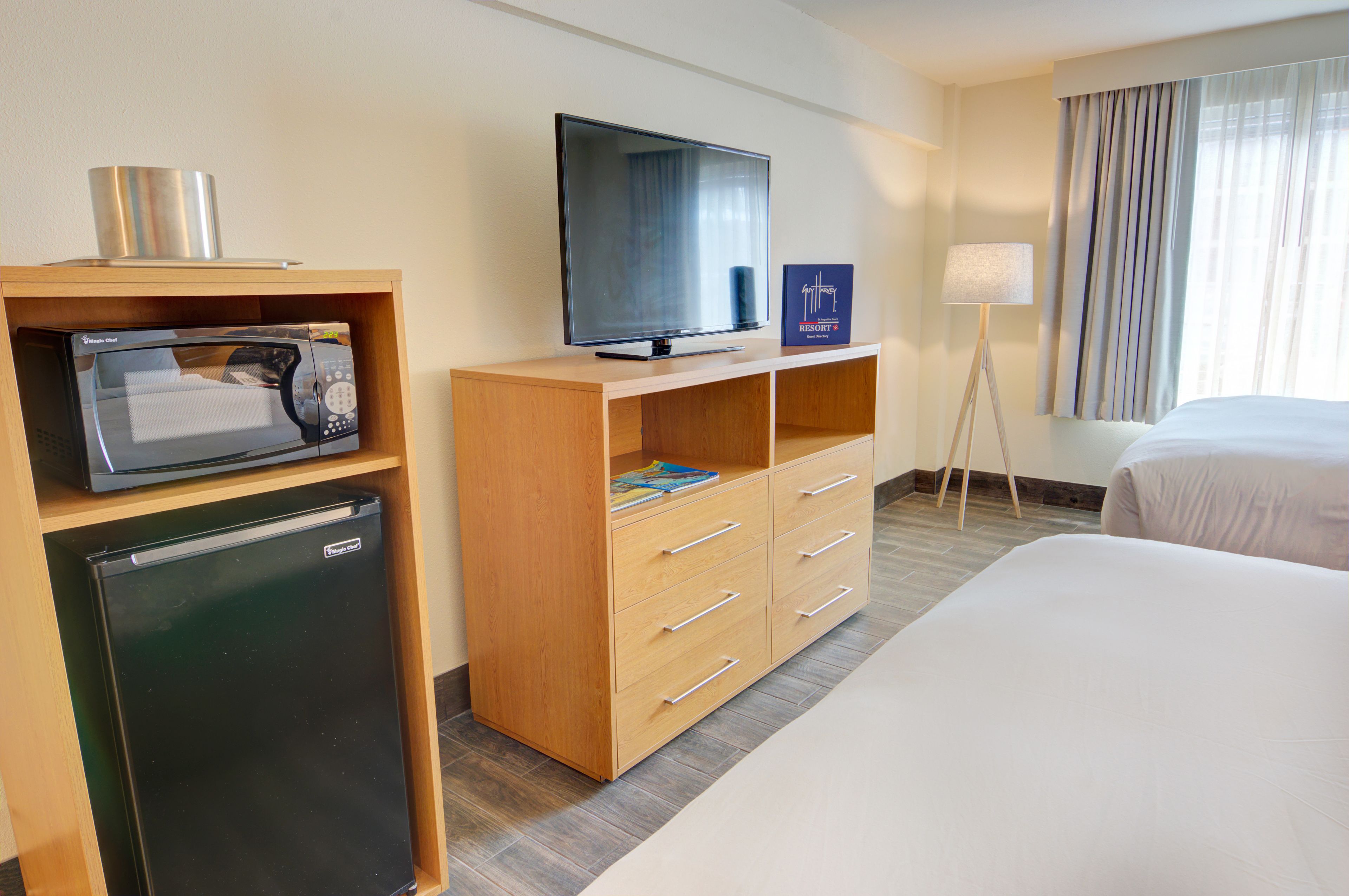 A hotel room with a bed, a TV, a mini-fridge, a microwave, a lamp, and a window with curtains.